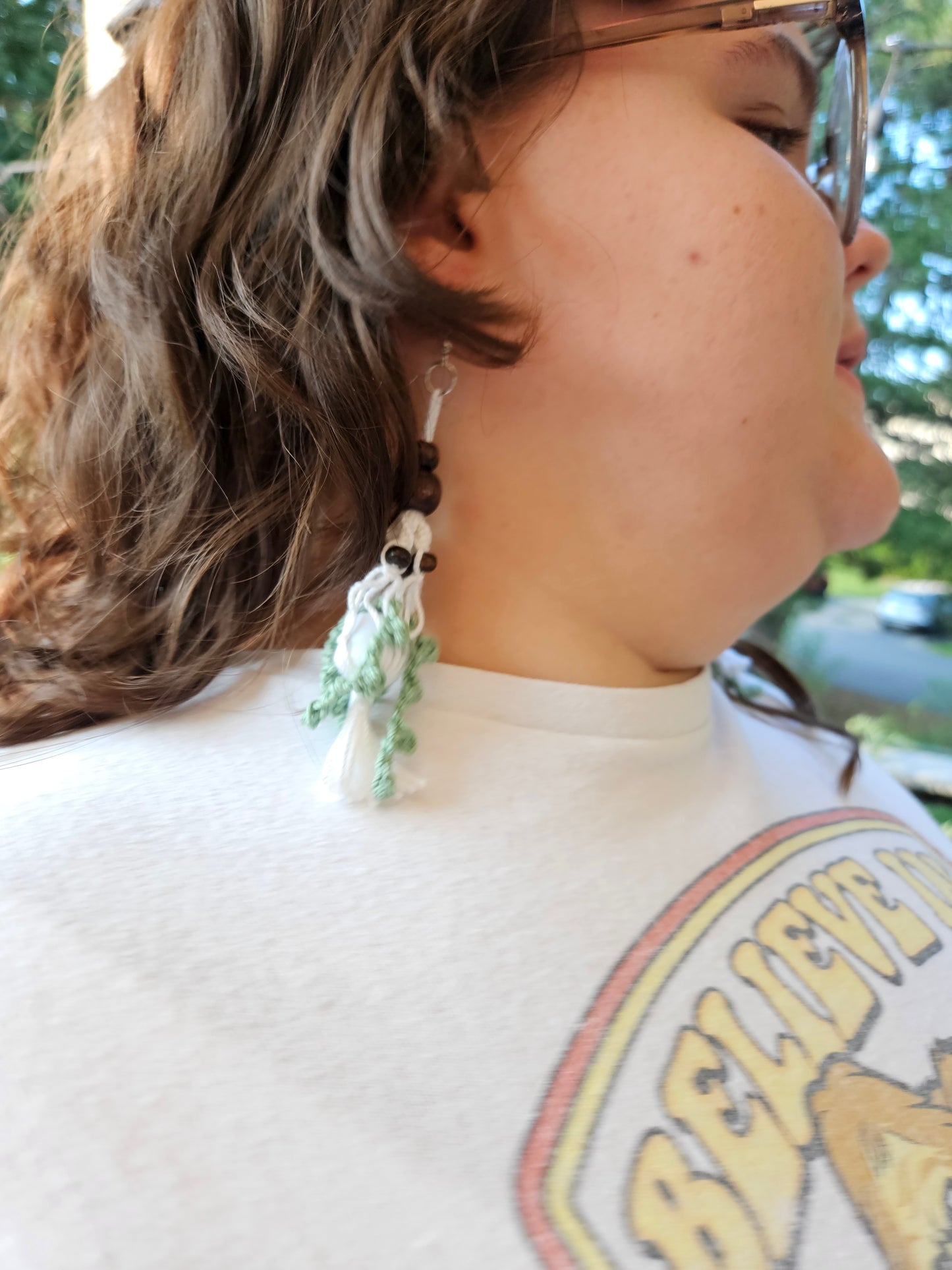 Mini Crochet Hanging Plant Dangle Earrings - Vine and Dark Wood Beads Right earring modeled by a girl. Earring with green crochet vines in a grey pot. Hanging in a white macrame hanger adorned with dark brown wooden beads.