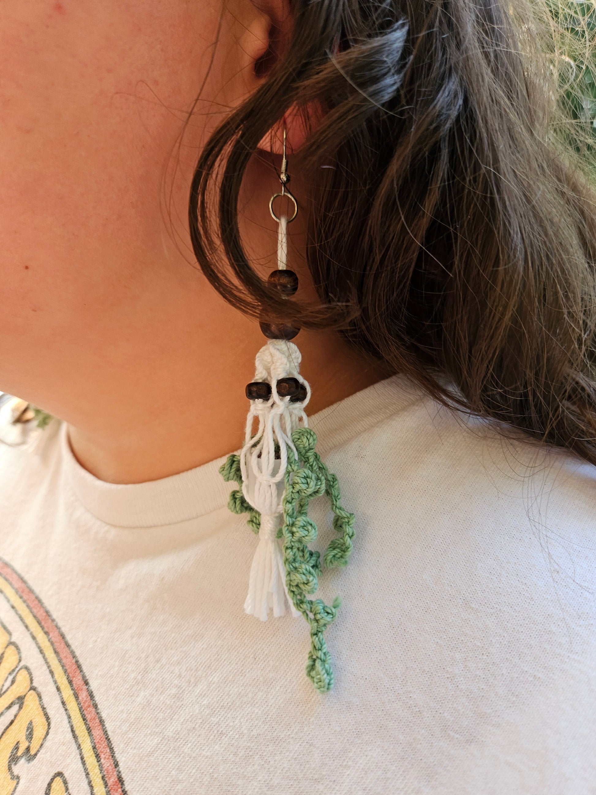 Mini Crochet Hanging Plant Dangle Earrings - Vine and Dark Wood Beads Left earring modeled by a girl. Earring with green crochet vines in a grey pot. Hanging in a white macrame hanger adorned with dark brown wooden beads.