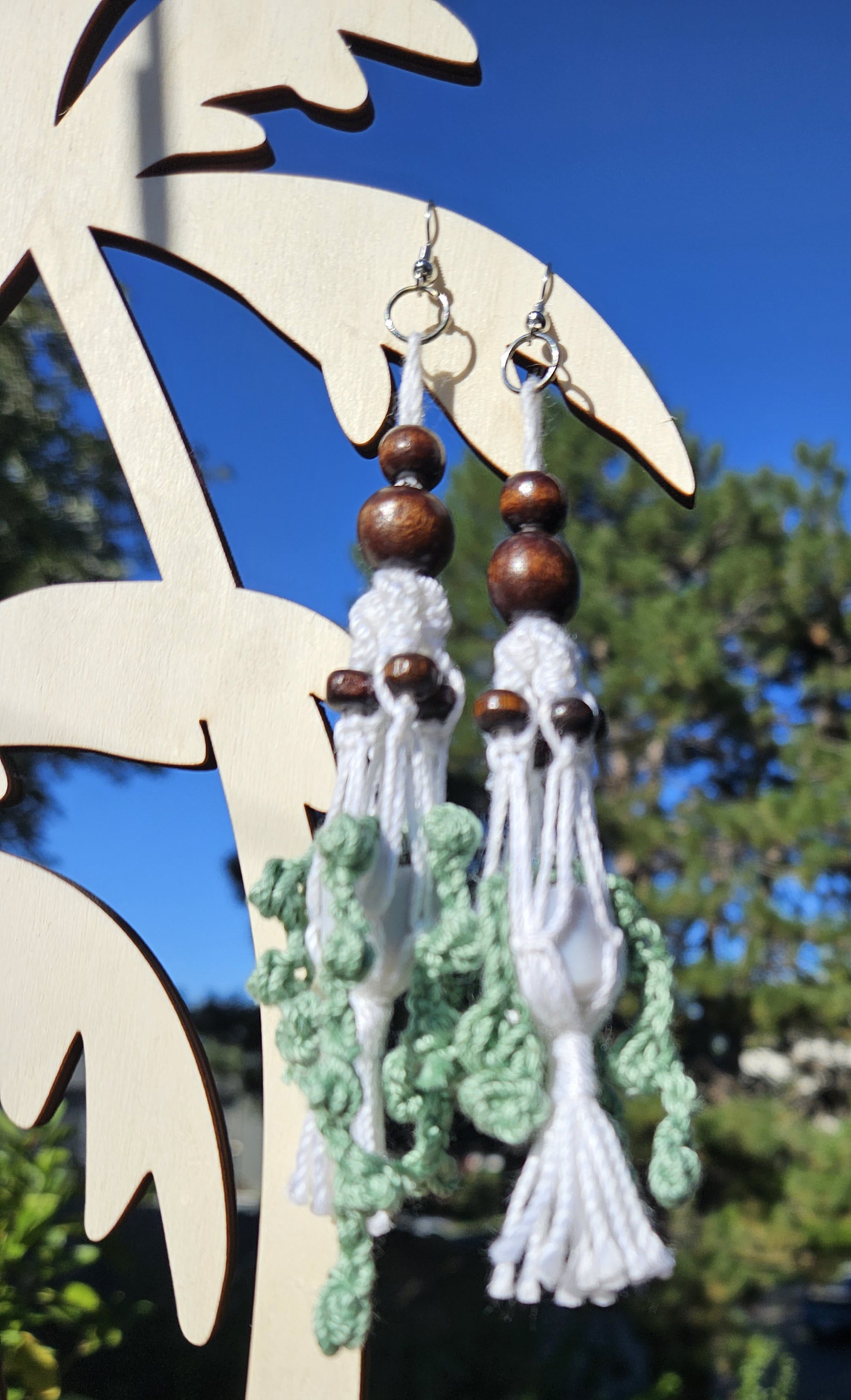 Mini Crochet Hanging Plant Dangle Earrings - Vine and Dark Wood Beads Pair of hanging plant earrings with green crochet vines in a grey pot. Hanging in a white macrame hanger adorned with dark brown wooden beads.